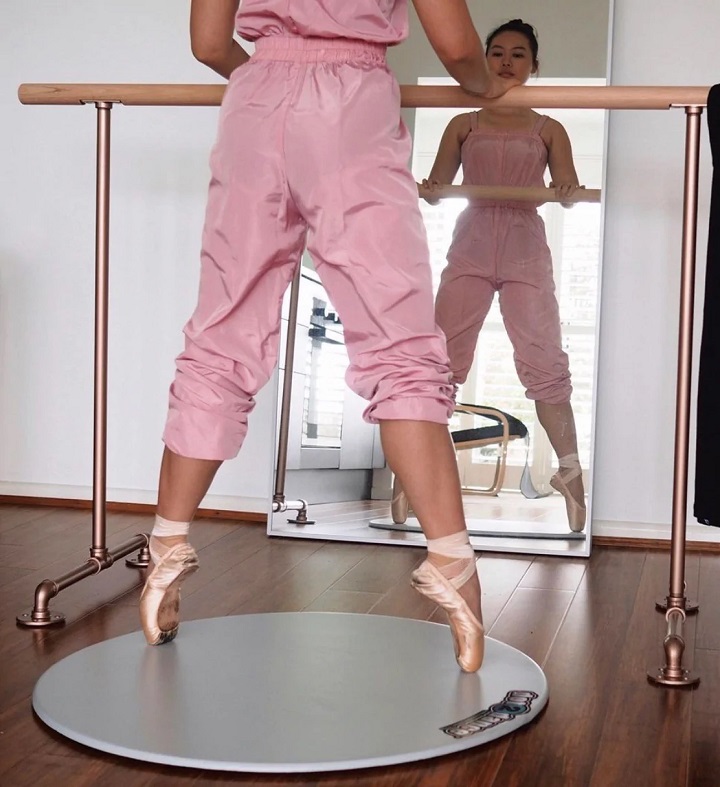 picture of a ballerina in a dance studio in front a mirror holding herself to a ballet barre standing on a turning board for ballet foot strecher