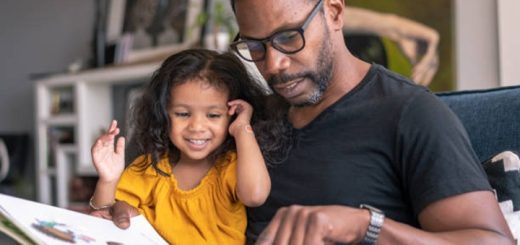 A loving father of African descent sits on the couch at home and reads a storybook to his preschool age daughter. The child is sitting on her father's lap and is smiling while looking at the book.
